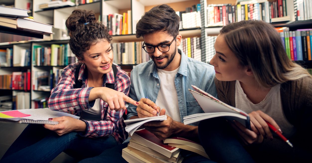 Close up view of three attractive beautiful cute stylish hipster young students learning together and sitting on the floor with a stack of book in the library.